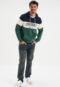 Uomo in felpa tricolore con il testo "CREEKS", jeans blu e scarpe bianche, si trova con le mani in tasca su uno sfondo bianco.
