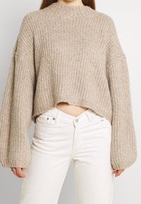 Beige stickad tröja med en kortad design, breda ärmar och ribbad textur, parat med vita högmidjade jeans.
