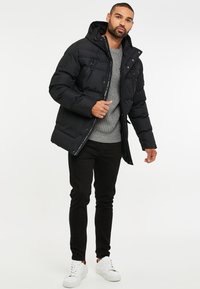 Chaqueta negra acolchada con capucha, que presenta dos bolsillos frontales con cremallera y un suéter gris texturizado debajo, combinado con pantalones negros ajustados y zapatillas blancas.