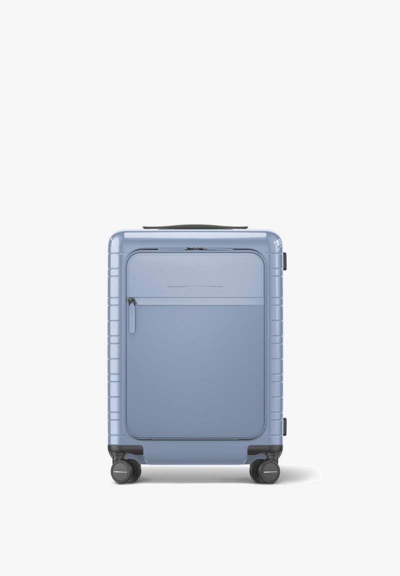 Hard-shell suitcase in light blue, featuring a front zipper pocket, black wheels, and a textured surface with horizontal grooves.