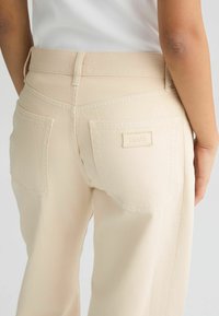 Pantalones de mezclilla beige claro con cintura alta, que cuentan con dos bolsillos traseros y una pequeña etiqueta de marca en el bolsillo derecho. Textura suave.
