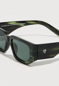 Πράσινα γυαλιά ηλίου με γωνιώδη σκελετό, υφασματικό μοτίβο, γκρι φακούς και λεπτομέρεια λογότυπου στο πλάι. Σχεδιασμένα για προστασία από τον ήλιο.