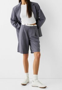 Šedé šortky na míru s záhyby, kombinované s bílým crop topem a šedým sakem, nošeny s bílými teniskami a ponožkami do lodiček. Hladká tkanina.