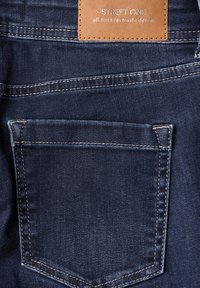 Tissu en denim bleu foncé, avec une poche arrière à couture contrastante et une étiquette en cuir marron embossée avec le nom de la marque.
