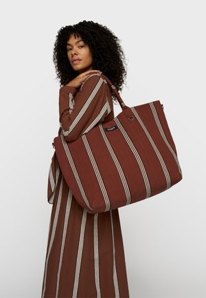 Kobieta o kręconych włosach, ubrana w brązową sukienkę w paski, niosąca na ramieniu dużą brązową torbę na ramię w paski na jednolitym tle.