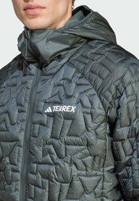Dunkelgrüne, gesteppte, isolierte Jacke mit Kapuze; ausgestattet mit einem durchgehenden Reißverschluss und dem 'TERREX'-Logo in Weiß auf der Brust. Glattes, leichtes Material.
