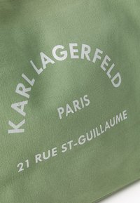 Sac en tissu vert avec un texte blanc en relief : "KARL LAGERFELD PARIS 21 RUE ST-GUILLAUME" et une surface texturée.