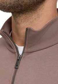 Veste marron à col zippé présentant un tissu texturé et des détails de fermeture éclair contrastants, étroitement ajustée autour de la zone du cou.