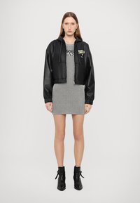 Schwarze Leder-Cropped-Jacke mit Kapuze, kombiniert mit einem gestreiften grauen Kleid. Das Model trägt schwarze Knöchelstiefel; Vorderansicht, neutraler Hintergrund.
