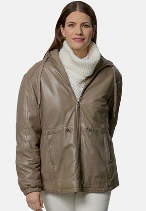 Woman wearing a taupe leather zip-up jacket over a white knit turtleneck sweater, standing against a plain background.