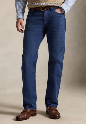 Un modèle cadré en partie porte un pantalon bleu foncé à jambes droites, des mocassins marron, un pull à motifs, une chemise rayée, une ceinture et une montre-bracelet.