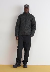 Melnas kviltotas jakas ar augstu apkakli un rāvējslēdzēju, apvienota ar melnām kravbiksēm, kurām ir kabatas. Melnas kurpes un pelēks cepure pabeidz šo tērpu.