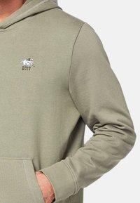 Jasnooliwkowa bluza z kapturkiem wykonana z miękkiego materiału, z kieszenią kangurką, ściągaczami przy rękawach oraz małym, białym haftowanym logo na piersiach.