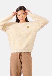 Maglione fluffy color crema con maniche lunghe e colletto rotondo, con un piccolo cuore dorato dettaglio sul petto. Indossato con pantaloni marroni brillanti.