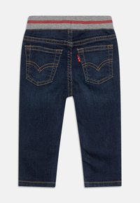 Jeans en denim bleu foncé avec une ceinture élastique grise comportant une bande rouge. Comprend des poches arrière doubles avec des détails de surpiqûre contrastés.