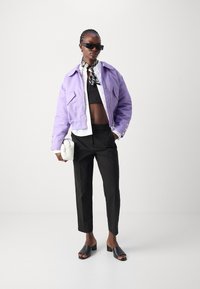 Levandulová kratší bunda s detaily na knoflících, černý crop top, upravené černé kalhoty, černé otevřené mule a bílá kabelka.