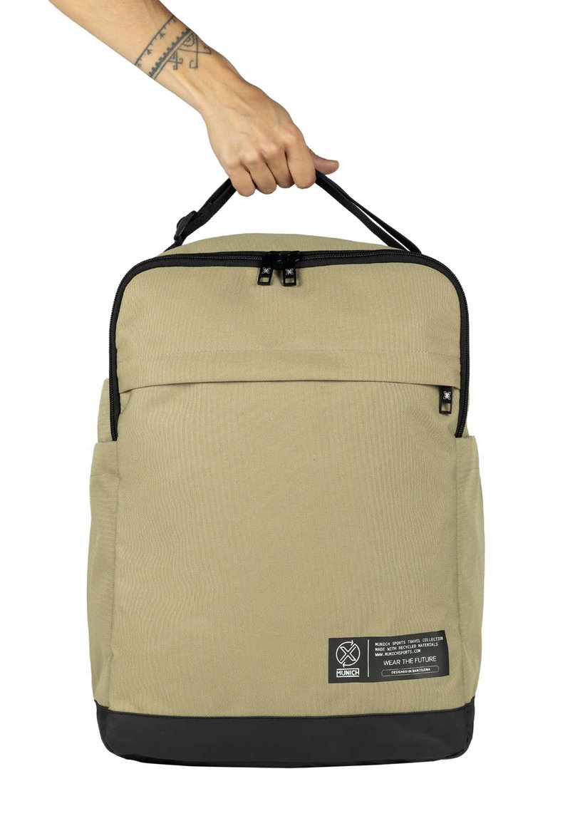 Sac à dos beige en tissu avec une finition texturée, doté d'un dessus zippé noir, d'une poche avant, d'une fente latérale et d'une étiquette en bas.