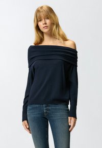 Mornarsko plavi džemper s off-the-shoulder dizajnom, rebrastim detaljima, mekom teksturom, dugim rukavima i opuštenim krojem, upairan s plavim trapericama.