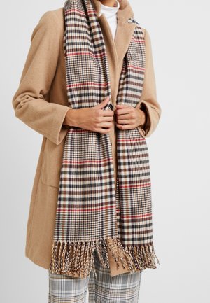 Personne portant un manteau camel et une écharpe à carreaux avec franges, assortie à un pantalon à carreaux, mains tenant l'écharpe devant.