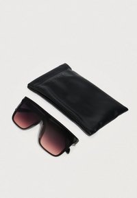 Μεγαλόσωμα μαύρα γυαλιά ηλίου με ροζ φακούς δίπλα σε μαλακή μαύρη θήκη προστασίας σε άσπρη επιφάνεια.