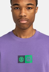 Purple sweatshirt featuring a crew neckline and ribbed cuffs, with a front graphic of green and black square logos. Soft texture.