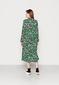 Vestido midi floral verde com mangas longas, apresentando flores brancas e roxas. Usado com ténis preto e branco. Tecido suave, ajuste relaxado.