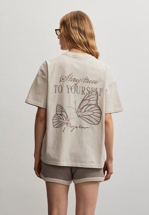 Kvinde med briller iført en beige T-shirt med teksten "Forbliv tro mod dig selv" og sommerfugleillustrationer, kombineret med strikkede shorts.