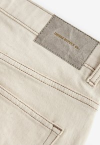Bež denim tkanina s vidljivim šavovima. Na stražnjoj strani se nalazi teksturirana siva oznaka utisnuta s natpisom "DENIM SUPPLY Co."