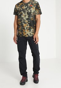 T-shirt camouflage vert, marron et noir, associé à un pantalon cargo noir avec plusieurs poches, et des chaussures de randonnée noires robustes avec des accents rouges.