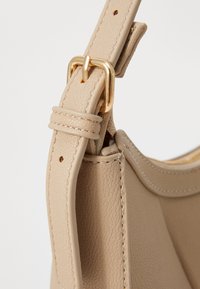 La sangle du sac à main en cuir beige possède une boucle en métal doré et des accents cousus, avec une surface texturée et une attache en boucle pour une longueur ajustable.