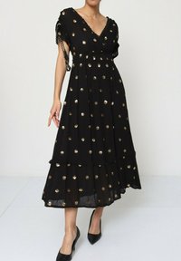 Robe maxi noire avec manches courtes, décolleté en V, motif à pois dorés, jupe à volants et détails aux épaules avec des accents en chaîne.
