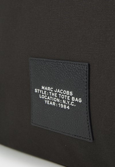 Borsa tote in tela nera con etichetta in pelle nera testurizzata con testo bianco in rilievo che indica dettagli di designer e stile.