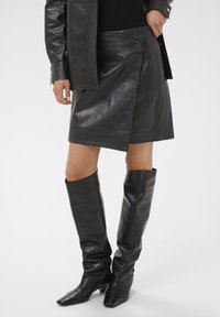 Falda envolvente de cuero negro con cierre de botones en la parte delantera y textura suave, combinada con botas de cuero negro hasta la rodilla con punteras puntiagudas.