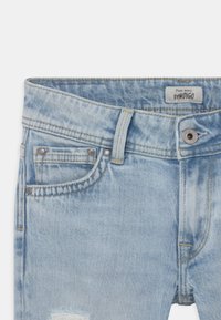 Jeans de mezclilla azul claro con un parche desgastado. Cuentan con cierre de botón y cremallera, presillas para cinturón y bolsillos delanteros con detalles metálicos.