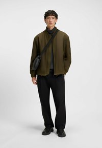 Olive green bomber jacket with ribbed collar, paired with a black textured sweater and loose black trousers. Black crossbody bag and shoes.