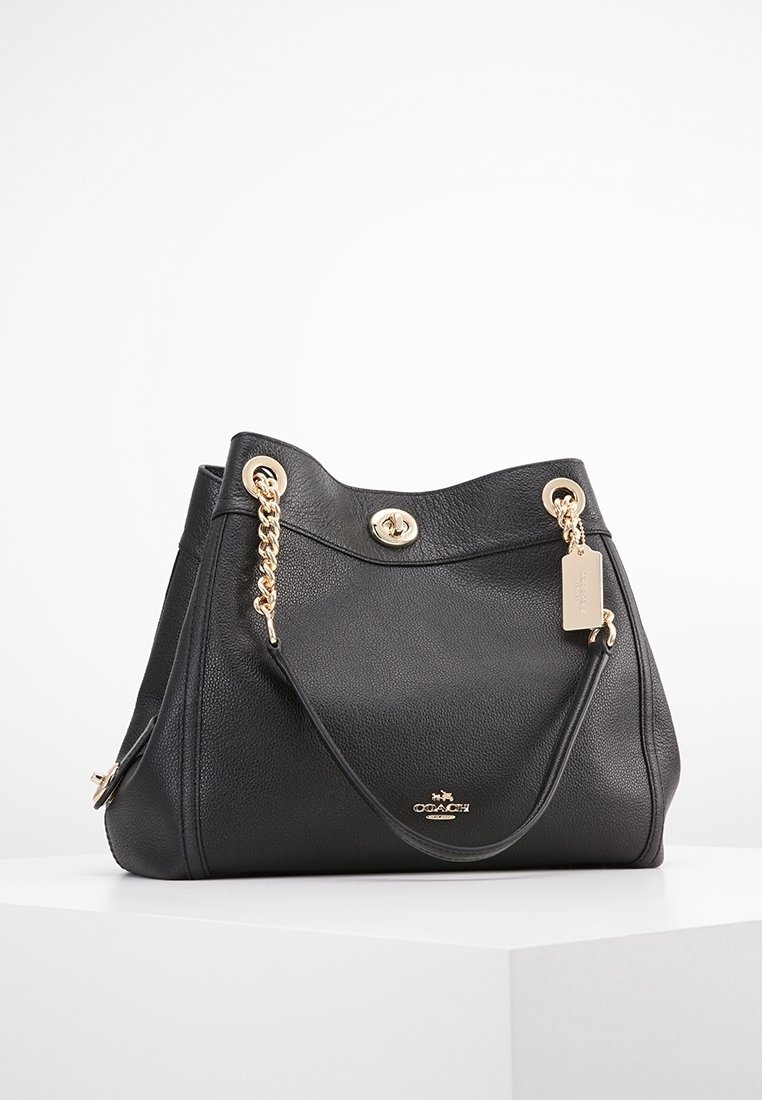Sac à main Coach en cuir noir avec des anses en chaîne dorée, fermeture à tourniquet et petit logo doré à l'avant, exposé sur un socle blanc.