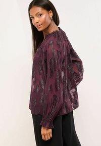 Sheer purple blouse with textured floral pattern, ruffled neckline, and elasticated cuffs, paired with black trousers.
