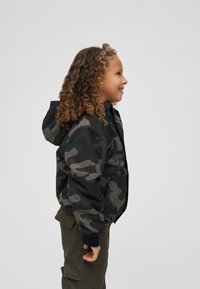 Veste camouflage noire et verte, avec capuche et poignets côtelés, associée à un pantalon cargo olive. Vue latérale du vêtement.