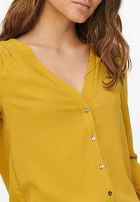 Blouse jaune avec un col en V, manches longues et devant boutonné. Texture douce, léger éclat et petits boutons argentés soulignant le design.