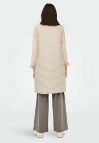 Kobieta ubrana w beżową, długą, pikowaną kamizelkę bez rękawów, jasną bluzkę, szerokie szare spodnie i beżowe buty, stojąca tyłem na jednolitym tle.