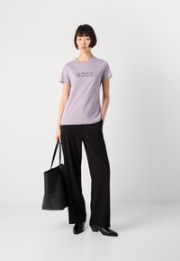 Moteris stovi, vilkinti levandų spalvos „BOSS“ marškinėlius, juodas plačias kelnes, juodus batus, vienoje rankoje laikydama didelę juodą rankinę.