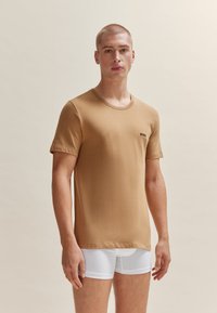 T-shirt brun à manches courtes en tissu lisse, avec un col rond et un petit logo noir sur la poitrine gauche. Porté avec des slips blancs.