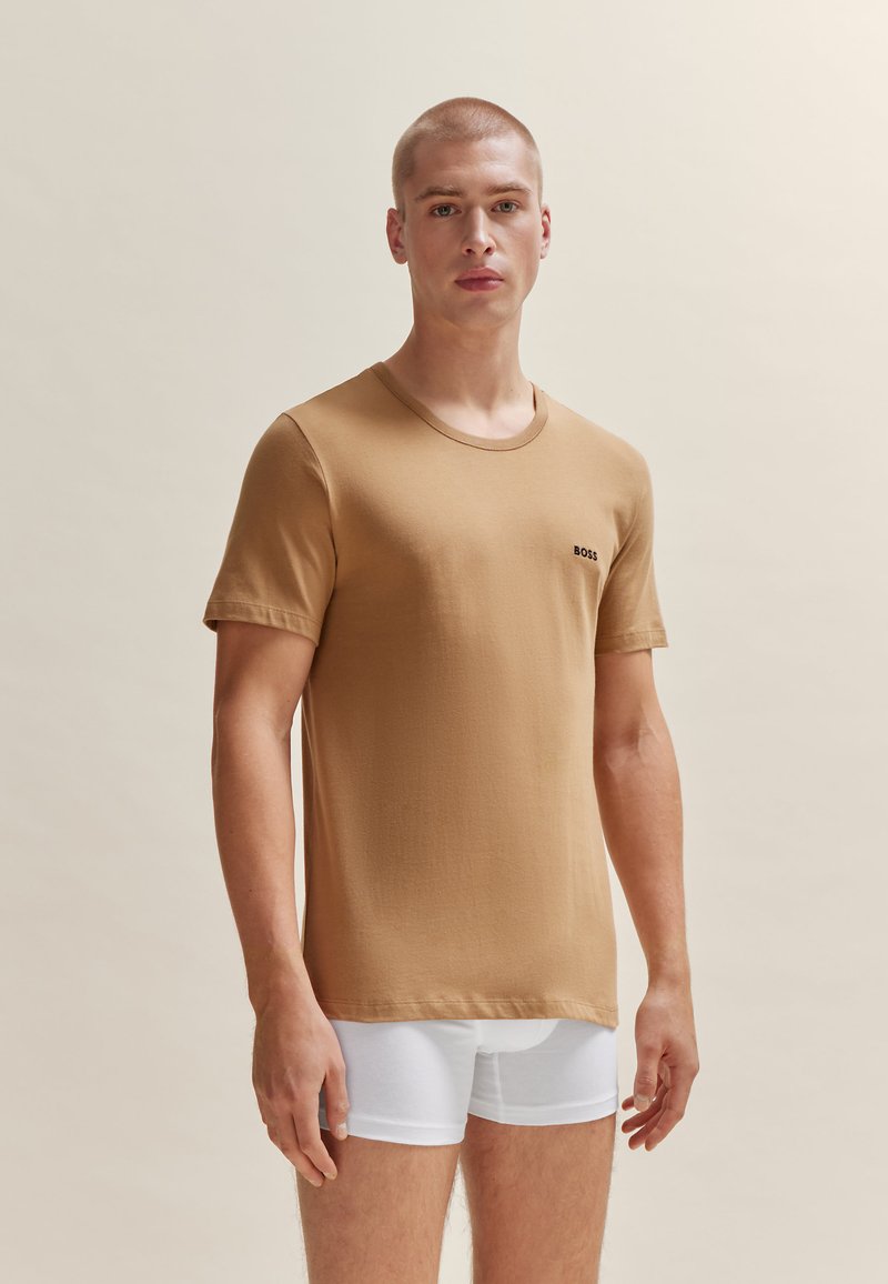 T-shirt brun à manches courtes en tissu lisse, avec un col rond et un petit logo noir sur la poitrine gauche. Porté avec des slips blancs.