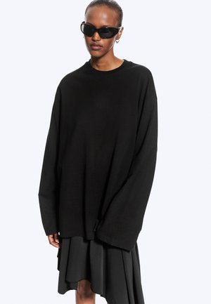 Personne portant une chemise noire oversize à manches longues, une jupe noire plissée, des lunettes de soleil noires et de petites boucles d'oreilles anneaux sur un fond clair.
