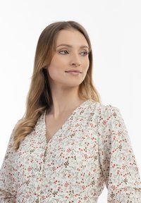 Bluse mit floralem Muster aus leichtem Stoff; zeigt rote und braune Blumen auf einem cremefarbenen Hintergrund, mit V-Ausschnitt und Knopfdetails.