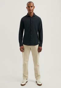 Camisa de manga larga gris oscuro con diseño de botones, combinada con pantalones beiges y zapatillas blancas, presenta un cuello clásico y un corte ajustado.