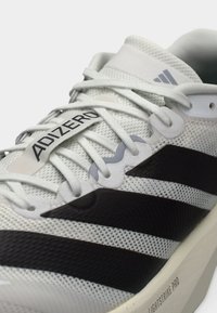 Weißer Sportschuh mit schwarzen Streifen, Mesh-Obermaterial, flachen Schnürsenkeln und einer hellgrauen Sohle mit der Aufschrift "LIGHTSTRIKE PRO" und "ADIZERO".