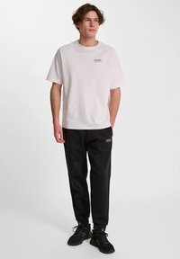 Ljusrosa T-shirt med rund halsringning och korta ärmar, tillsammans med svarta joggers med en logo och sportig design. Svarta sneakers.