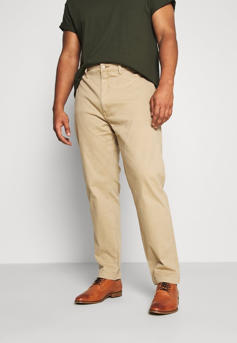 Chino beige in cotone con vestibilità dritta, indossati con una t-shirt a girocollo verde scuro e scarpe in pelle marrone. Presenta maniche arrotolate.