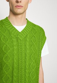 Gilet vert tricoté avec un col en V et des manches courtes, présentant un motif torsadé texturé. Porté par-dessus un t-shirt blanc.